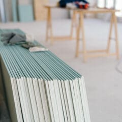 A stack of drywall panels leans against a wall in a bright, unfinished room, ready for the dry lining contractor. A pair of work gloves rests on top, with blurred work tables, tools, and equipment hinting at an office fit-out in progress in the background.