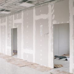 A corridor with unfinished drywall partitions reveals the meticulous work of a partitioning expert, showcasing taped seams and door openings. The floor is partially covered with sheets of cardboard, while the ceiling features a grid pattern complemented by some exposed wiring.