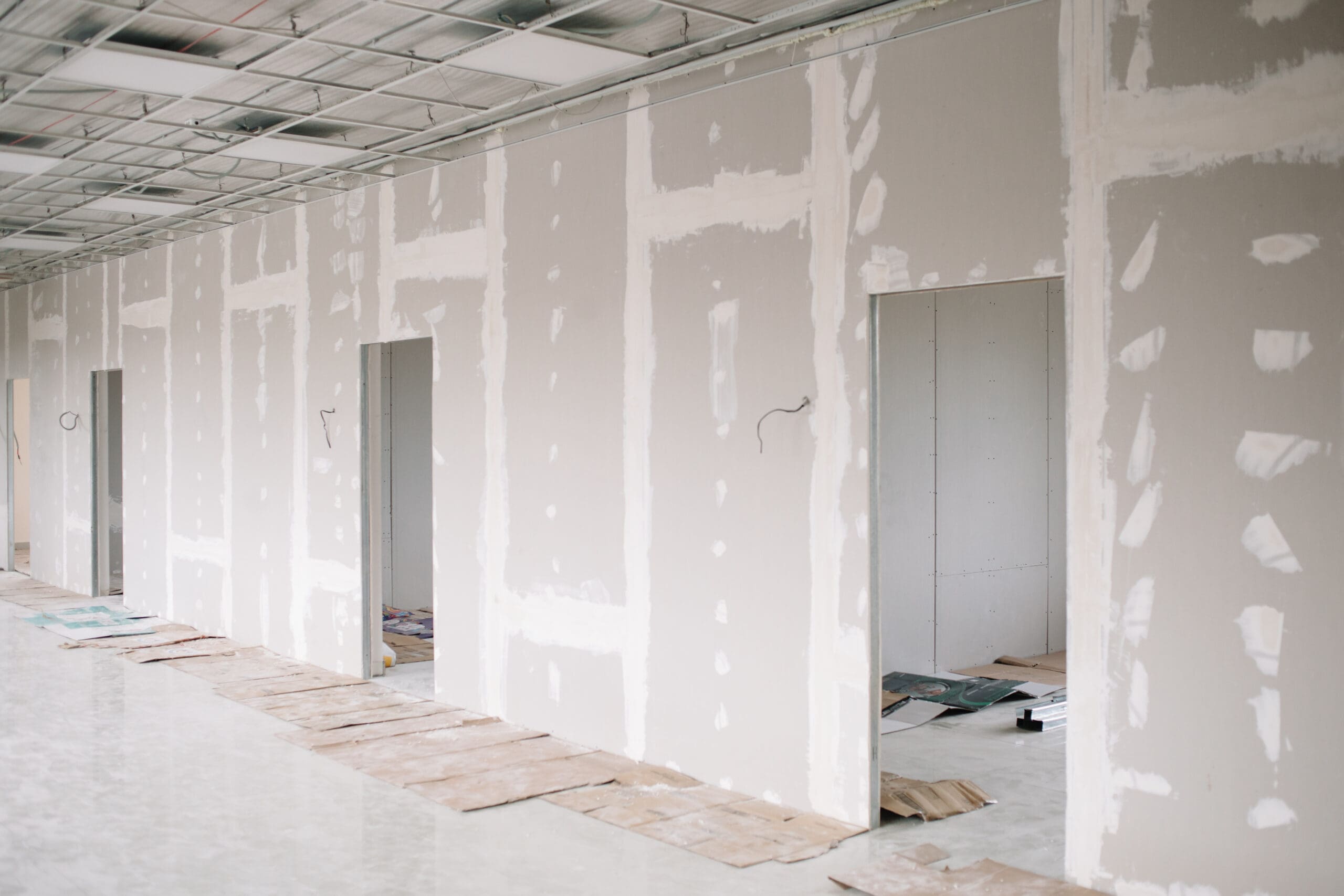A corridor with unfinished drywall partitions reveals the meticulous work of a partitioning expert, showcasing taped seams and door openings. The floor is partially covered with sheets of cardboard, while the ceiling features a grid pattern complemented by some exposed wiring.