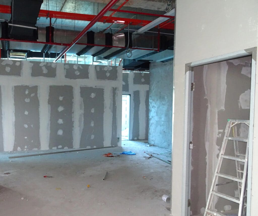 The interior of a building under construction showcases unfinished drywall by a skilled dry lining contractor, with visible joint compound. A metal ladder leans against the wall, and scattered construction materials clutter the bare concrete floor. Above, exposed ceiling pipes and ducts are visible.