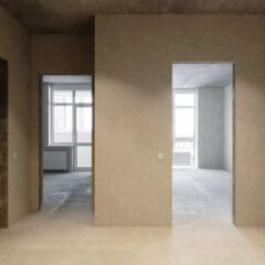 The minimalist interior of this concrete apartment boasts bare walls and floors, with two open doorways leading to rooms where large windows pour in natural light. The space is elegantly illuminated by recessed lighting, seamlessly integrated with suspended ceilings for a sleek touch.