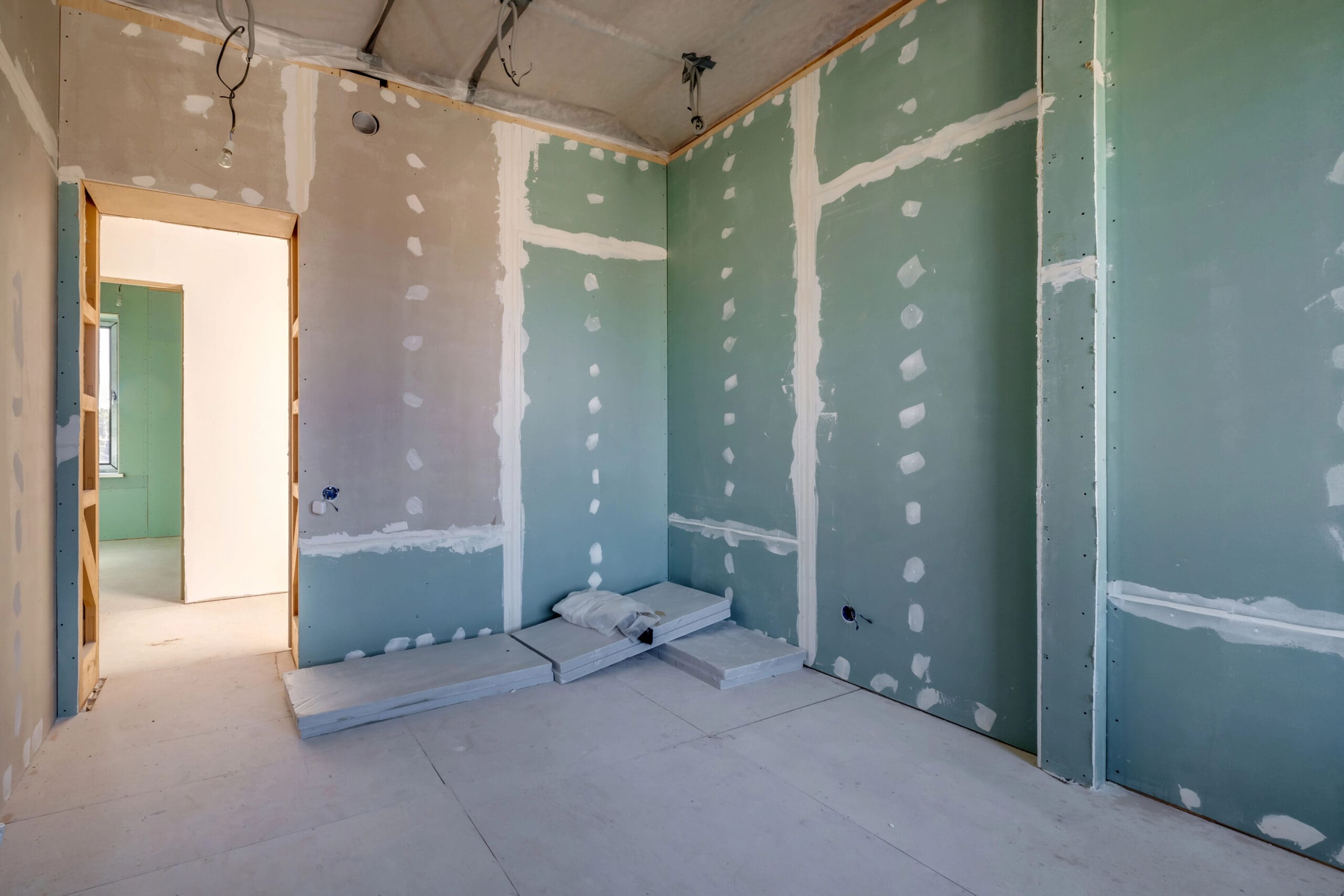 Room under construction with unfinished walls and exposed drywall. Green wall panels are partially finished with white joint compound by a skilled dry lining contractor. Uninstalled boards, essential for partitioning, are stacked on the floor. An open doorway leads to another room with light streaming in.