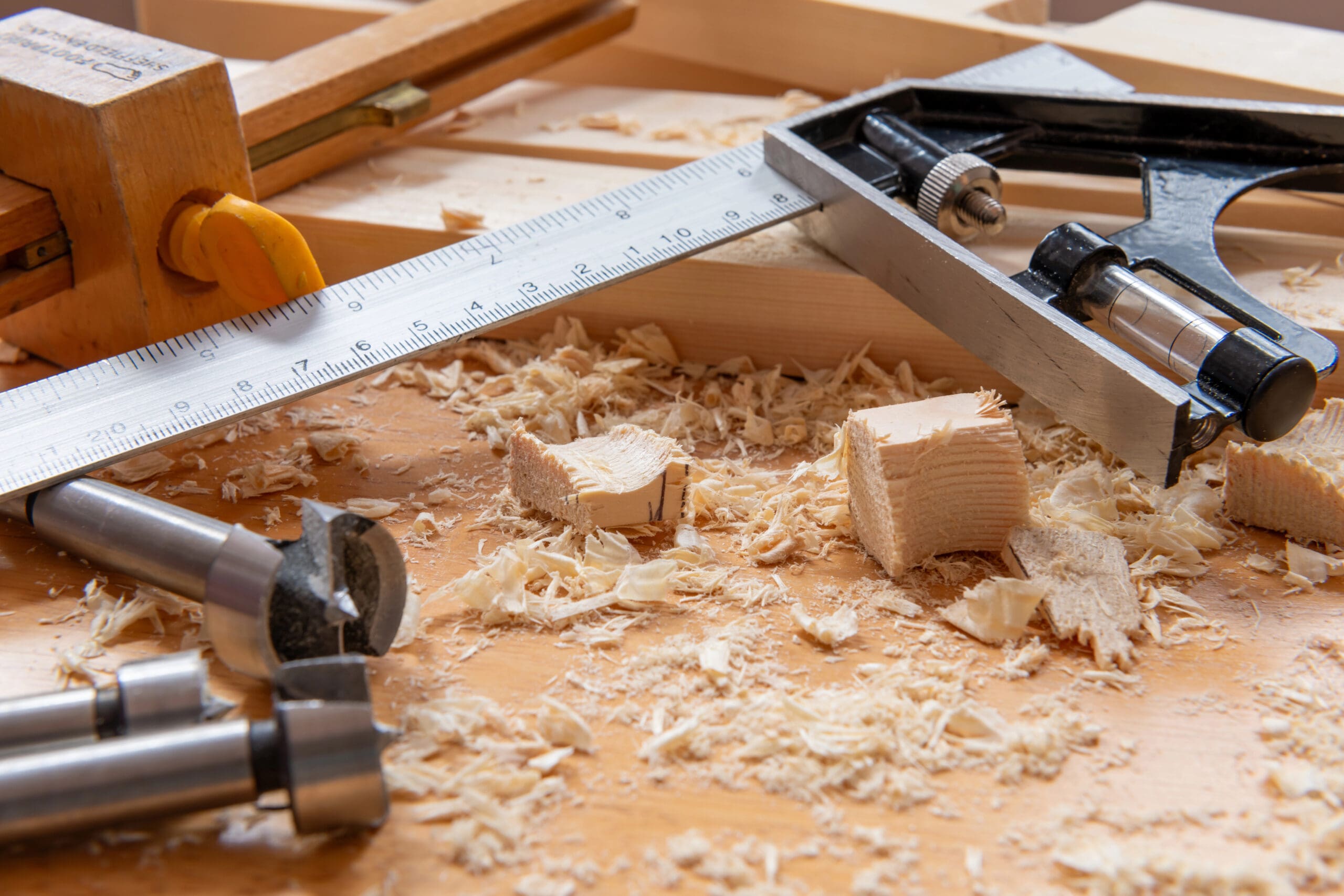 Woodworking tools on a workbench with wood shavings and hints of partitioning accessories. A metal square ruler, carpenter's square, drill bits, and a wooden vise are visible amid small wood chips and pieces.
