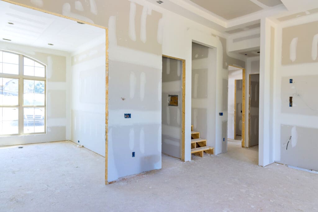 Unfinished interior of a house under construction, featuring drywall by a skilled dry lining contractor on walls and ceiling. Doorways lead to different rooms, and temporary wooden steps are visible. A large arched window allows natural light into the space, and the bare floors await finishing touches.