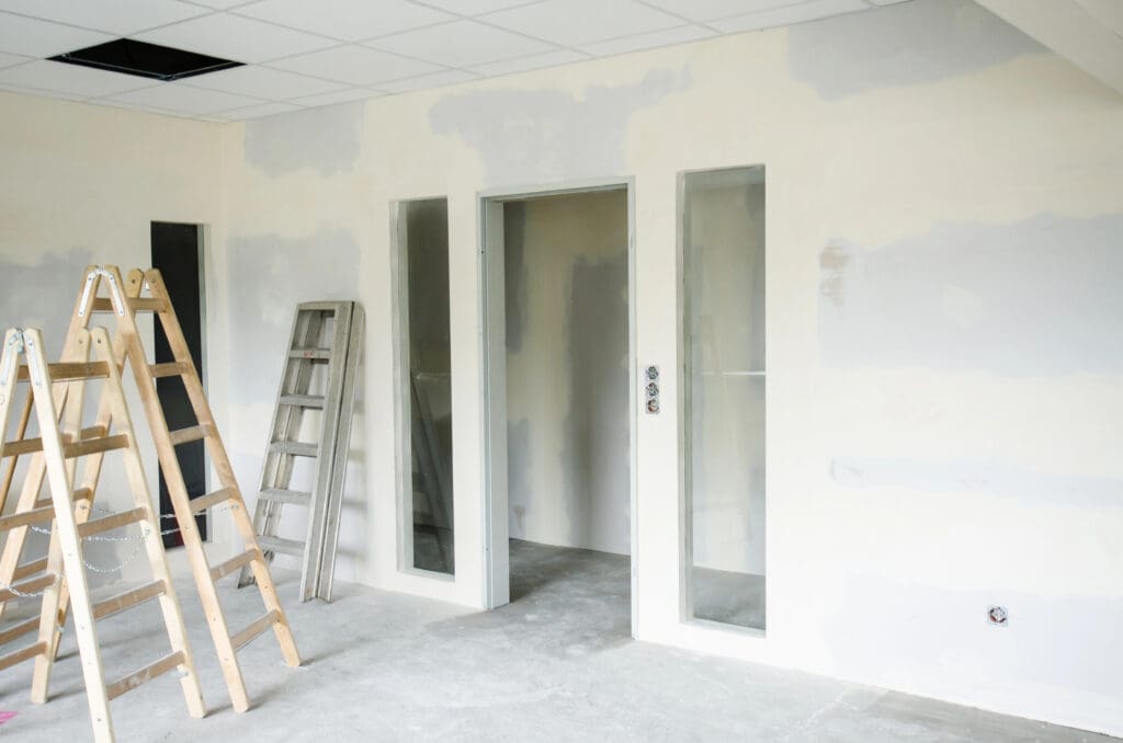 The room under renovation features a wooden step ladder and a metal ladder against the partially painted wall. At the center is a glass-paneled door, while sections of suspended ceilings reveal open tiles above. The bare concrete floor awaits its next phase in this office fit-out project.