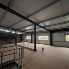 The interior of the unfinished building features a high ceiling with exposed beams, hinting at future suspended ceilings. Partitioning plans lie in wait as a ladder and bucket rest on the floor. Large windows along the far wall invite streams of natural light to fill this evolving space.