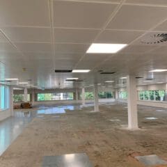 An empty, spacious office features suspended ceilings and support columns. Large windows flood the space with natural light. The floor is partially tiled, with some areas unfinished. A few cardboard boxes are stacked in the corner near the windows, awaiting carpentry work.