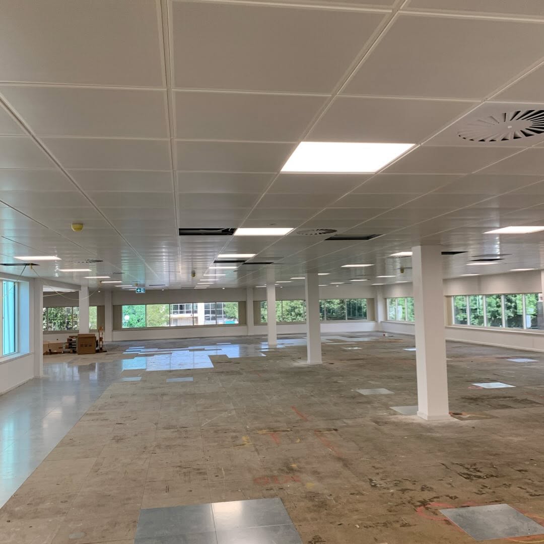 An empty, spacious office features suspended ceilings and support columns. Large windows flood the space with natural light. The floor is partially tiled, with some areas unfinished. A few cardboard boxes are stacked in the corner near the windows, awaiting carpentry work.