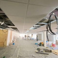 A large indoor construction site features a partially built ceiling with exposed wiring, indicating the work of a dry lining contractor. The floor is covered with protective sheets and tools, while workers are busy in the background. Daylight streams in through windows, illuminating the dynamic scene.