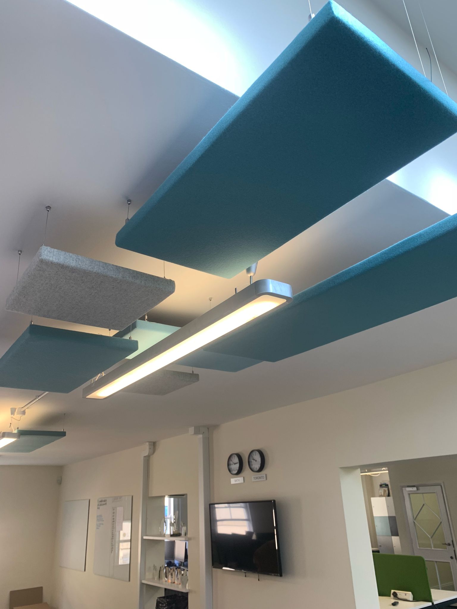 Ceiling view of the room reveals a suspended ceiling with rectangular acoustic panels in blue and gray, part of an office fit-out. A fluorescent light fixture complements this design. Below, a wall-mounted TV, whiteboard, and two clocks complete the space's functionality.