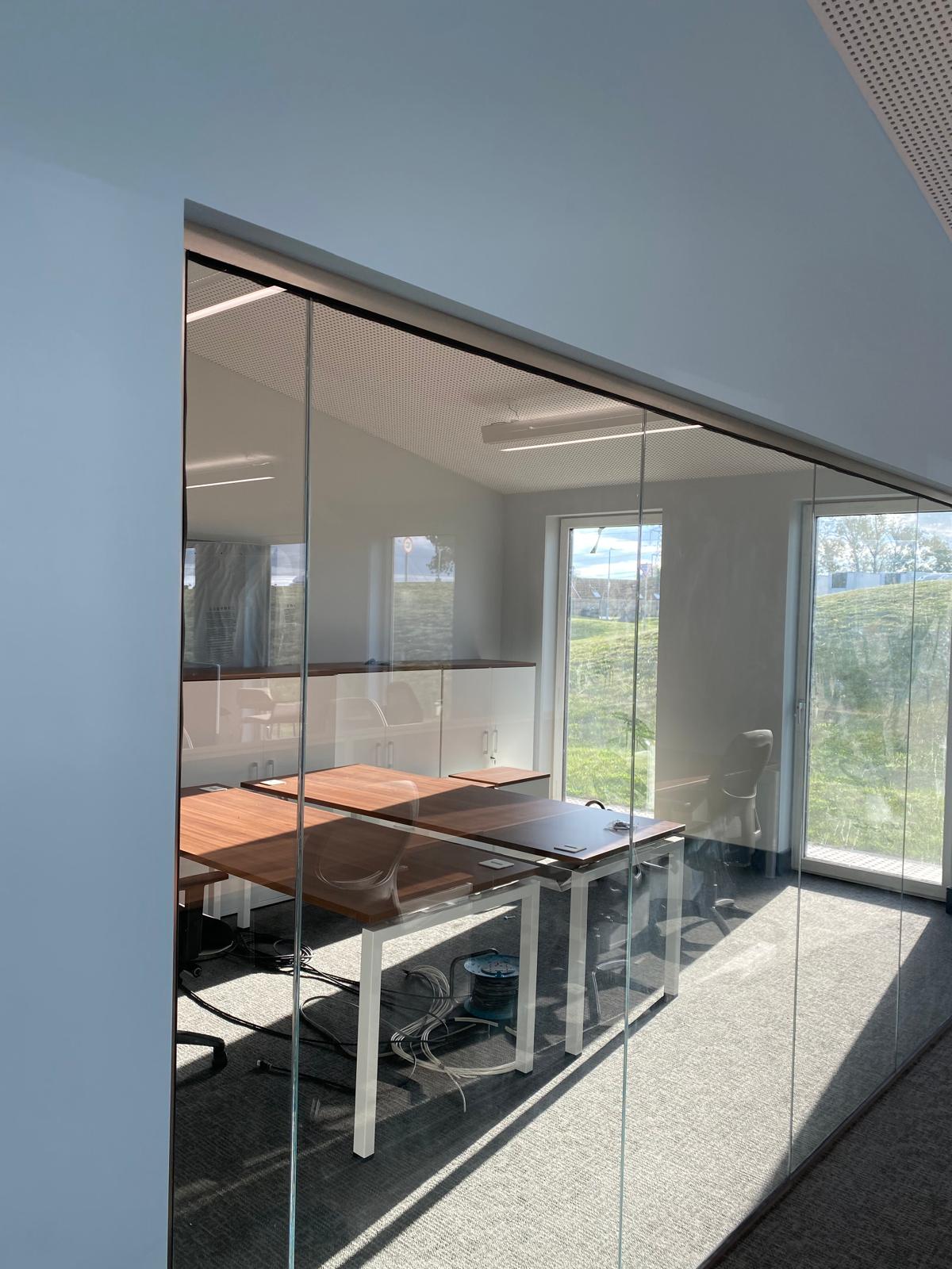 Glass-walled office with a wooden conference table and office chairs. Sunlight pours through large windows, highlighting the sleek partitioning and modern decor. The minimalistic space seamlessly blends with suspended ceilings, while a grassy landscape is visible outside.