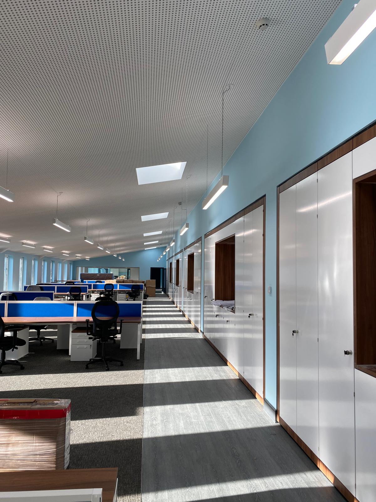 A modern office interior boasts a row of cubicles on the left and a hallway lined with closed cabinets on the right. The ceiling features recessed lights set in sleek suspended ceilings, while large windows allow natural light to cast intricate patterns on the floor.