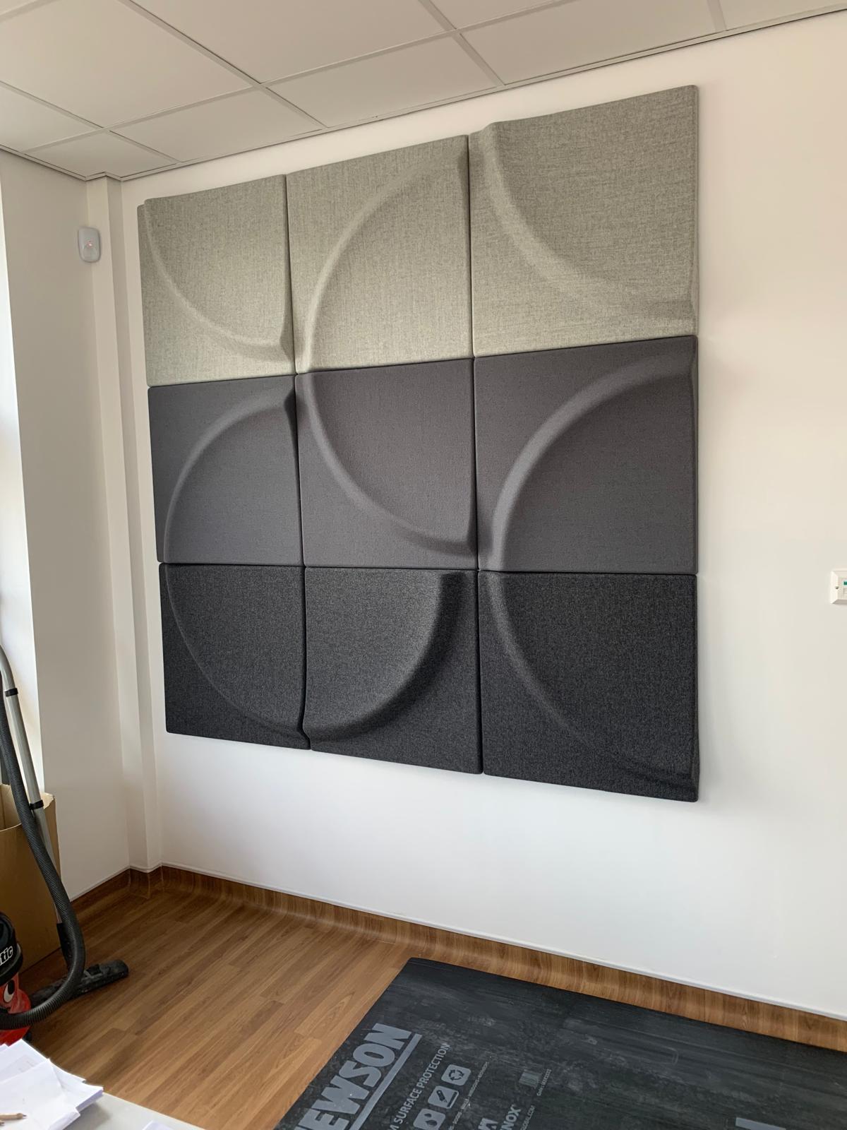 Wall-mounted acoustic panels, expertly crafted by a dry lining contractor, form a grid of nine with circular, abstract designs in varying shades of gray. The room's wooden flooring complements the subtle palette, while a partially visible treadmill rests to the left.
