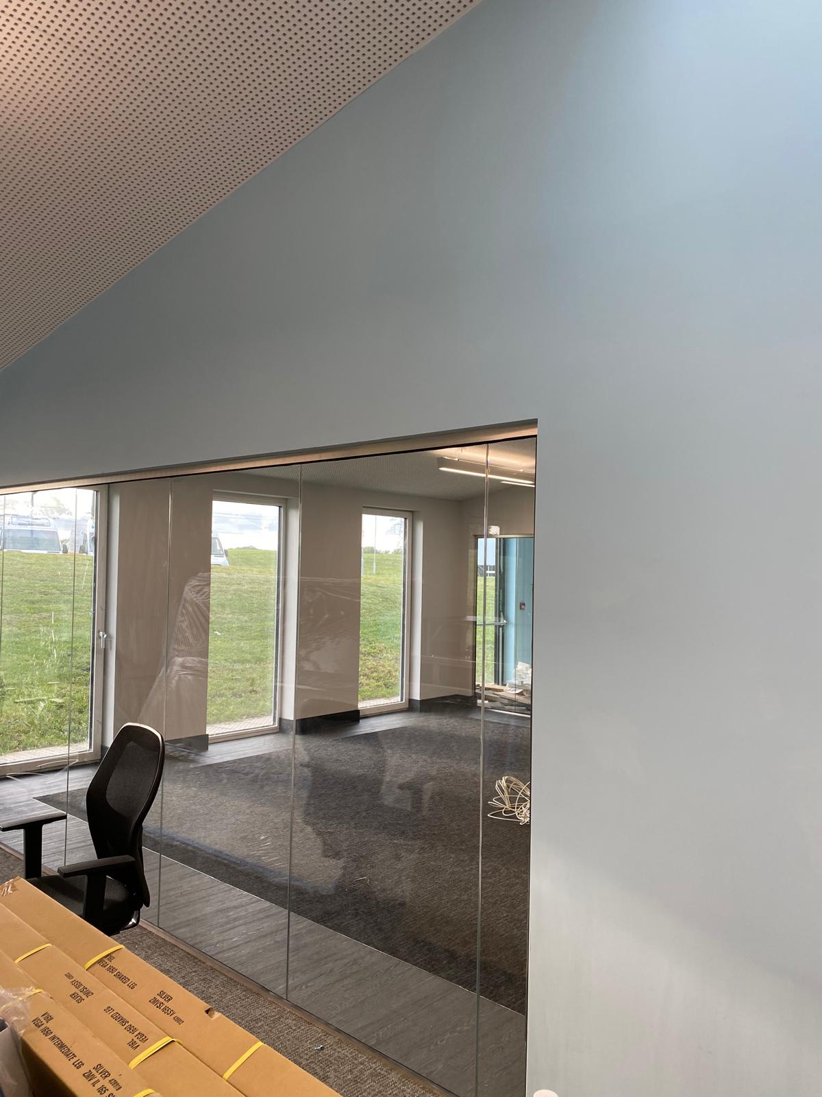 Office space with large windows framing a grassy outdoors. Inside, a black office chair complements the desk's edge. The walls are painted light blue under sleek suspended ceilings. Ceiling lights illuminate the gray-carpeted floor in this well-designed office fit-out.