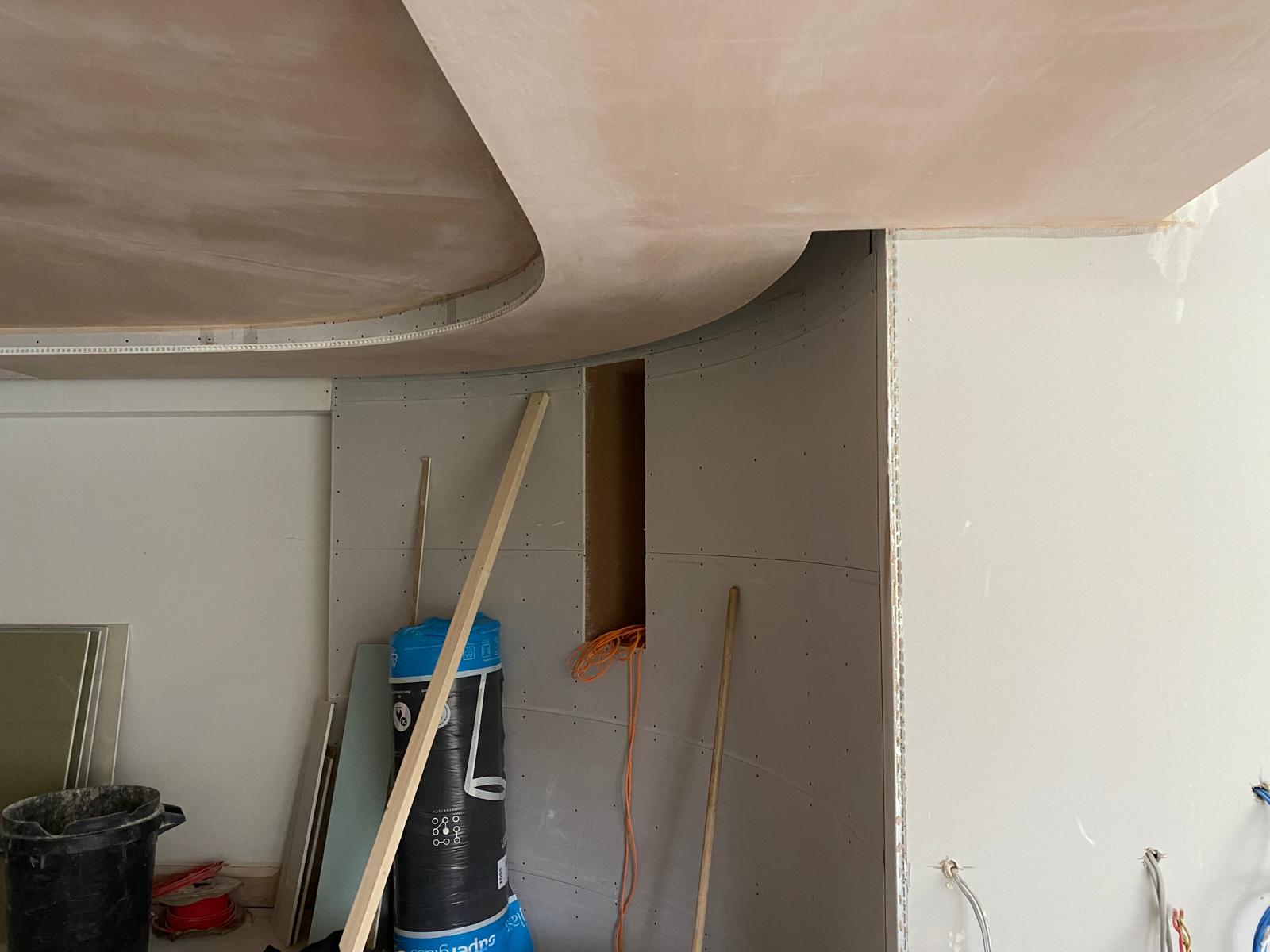 The room, part of an office fit-out project, features curved drywall and a covered cylinder amidst construction materials like a bucket and boards. An opening in the wall reveals cables, while a dry lining contractor works to complete the space with precision.