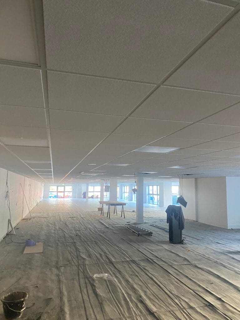 A large, empty room under construction with a grid ceiling and plastic-covered floor awaits office fit-out. Carpentry materials and worktables are scattered around as sunlight streams through windows at the far end, illuminating the space.