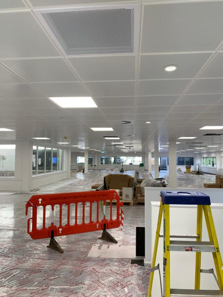 Modern office space under construction with ceiling lights and a red barrier. A ladder stands ready, hinting at meticulous carpentry work ahead. The floor is covered with protective plastic, while boxes and materials are scattered throughout, awaiting the dry lining contractor's skilled touch.