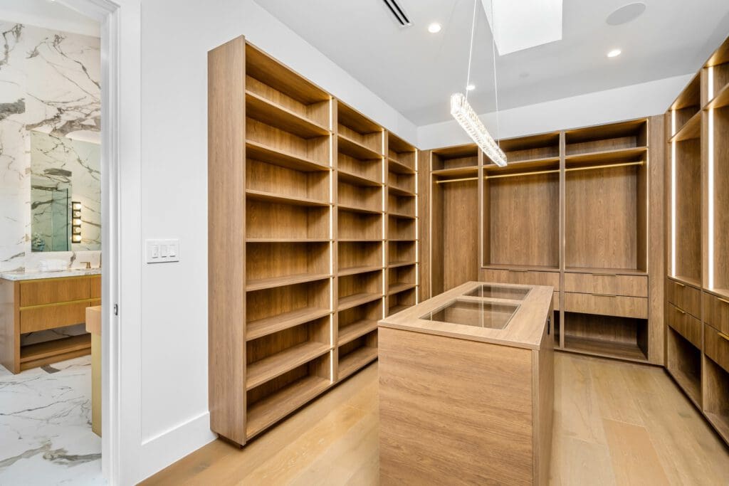 Spacious walk-in closet with wooden shelves and a glass-topped island. Features ample storage with floor-to-ceiling shelving and modern partitioning. A sleek light fixture hangs overhead, while in the background, a glimpse of a bathroom with marble accents is visible.