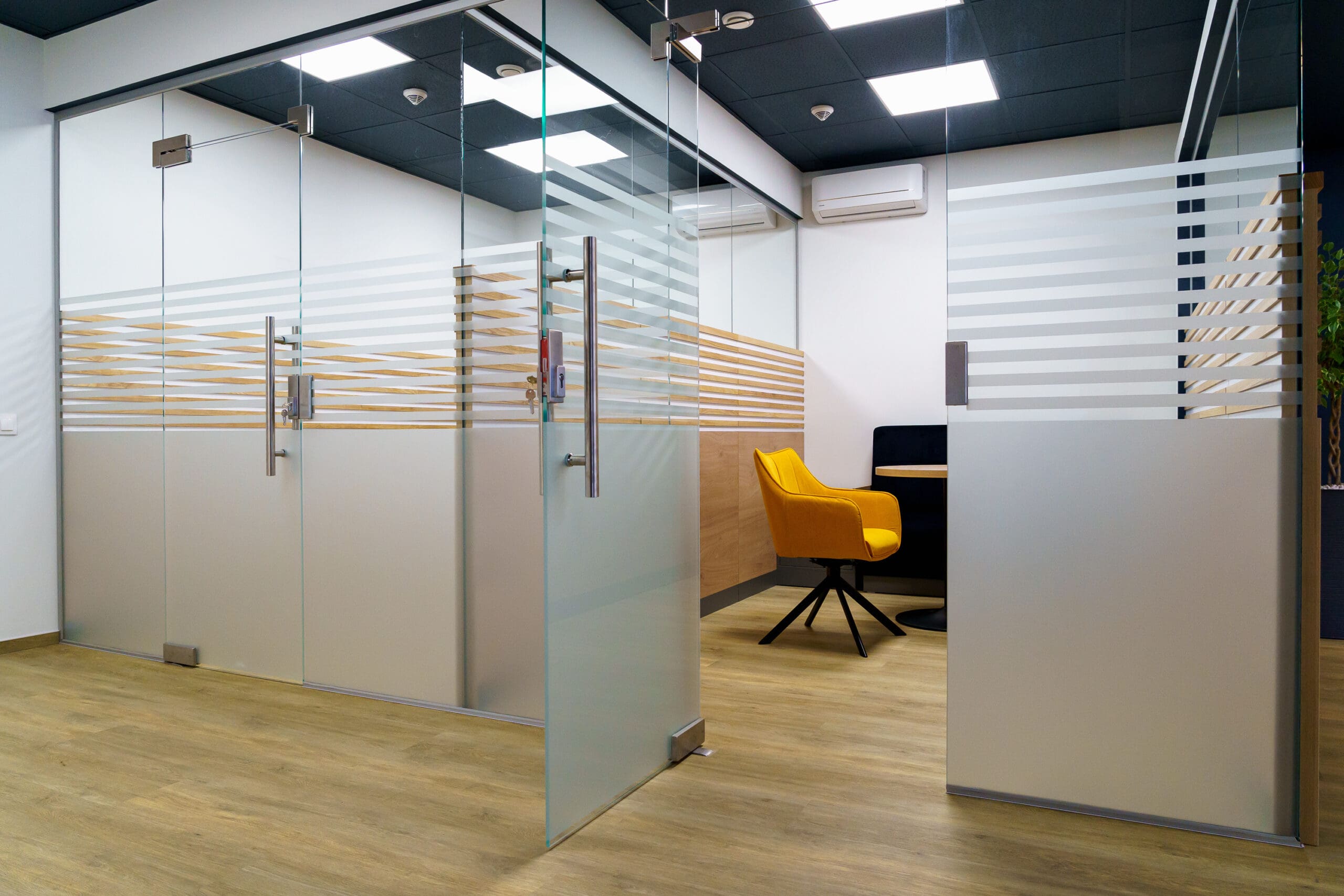 A modern office fit-out featuring glass partitioning separates rooms with frosted stripes. A closed room showcases a yellow chair and black table against a white wall, while wooden flooring and ceiling lights enhance the minimalistic design.