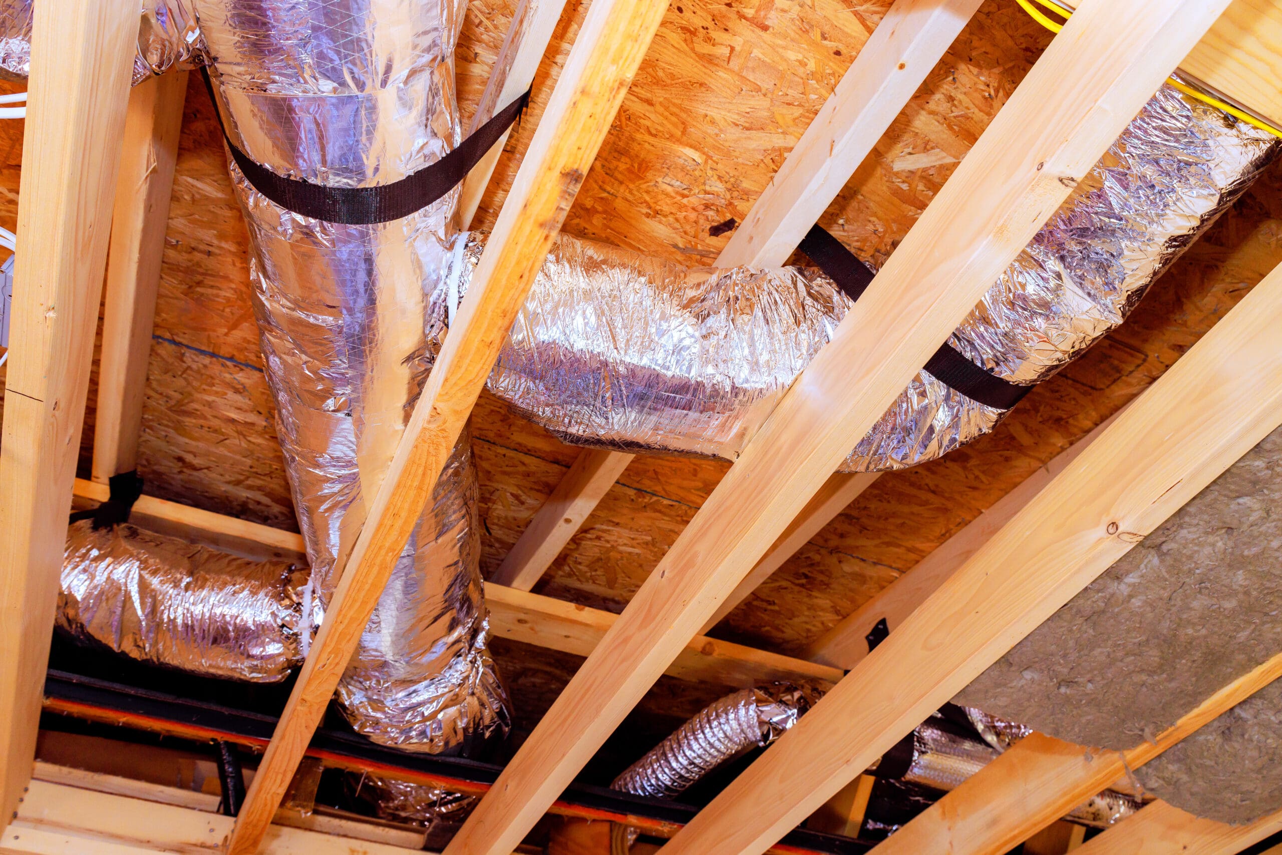 View of a ceiling with exposed wooden beams and shiny silver HVAC ductwork. The ducts, expertly handled by a dry lining contractor, are wrapped in insulation, running between the beams, and secured with black straps.