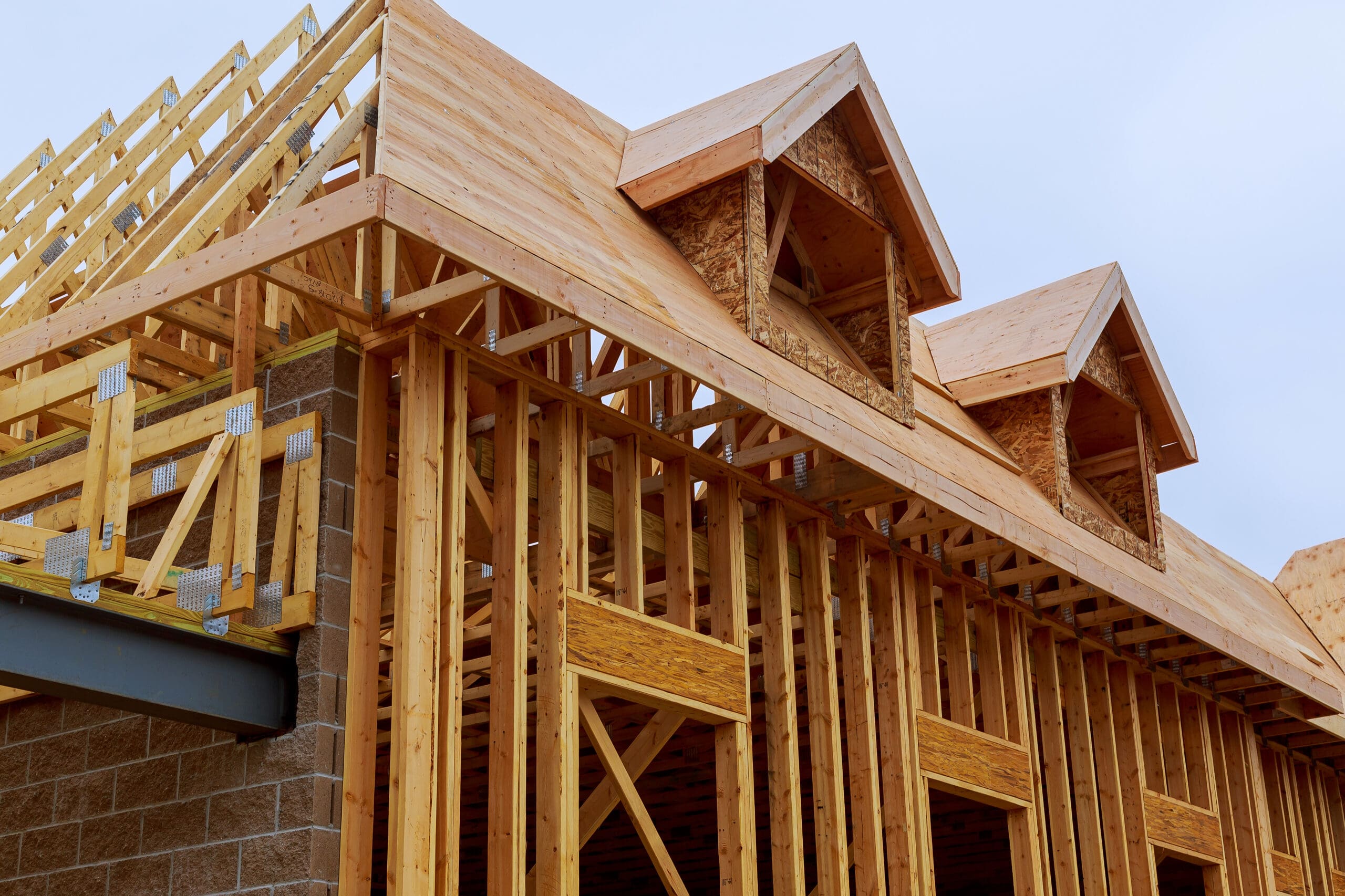 The wooden framework of a house under construction, showcasing expert carpentry with multiple dormer windows and a partially completed roof. The structure features various wooden beams and planks, set against a clear sky in the background.
