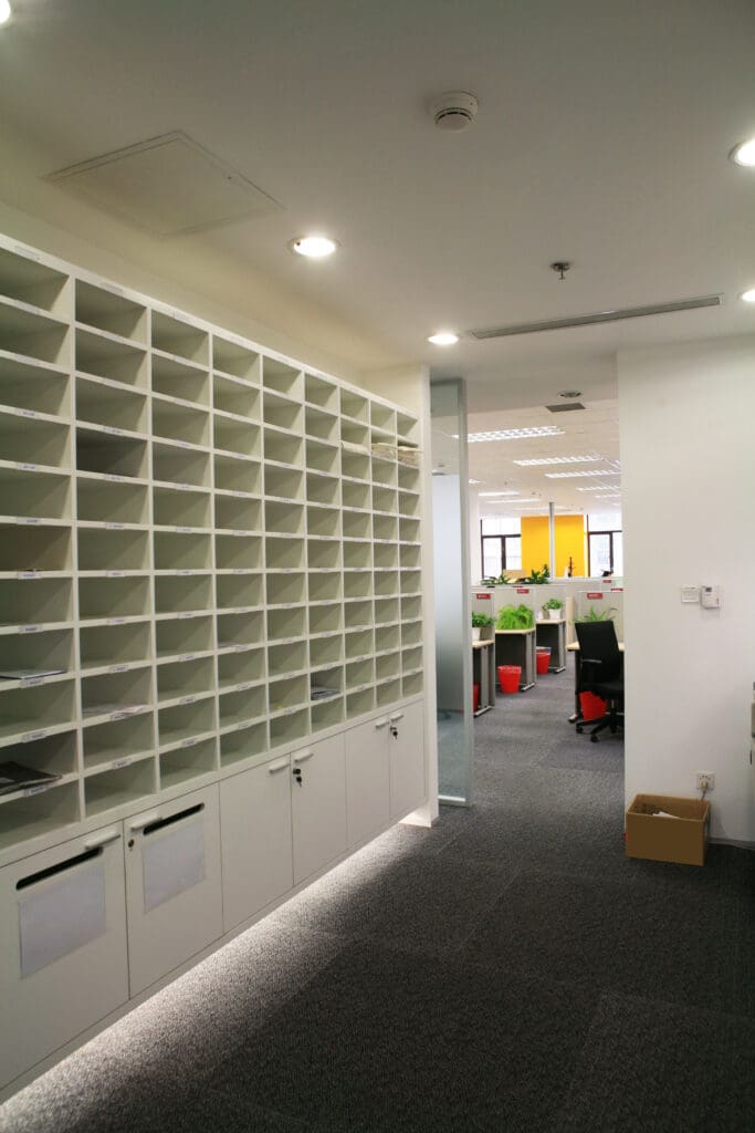 A modern office hallway showcases expert carpentry with a wall of white cubby shelves on the left, seamlessly leading to an open-plan office space. The area boasts desks and chairs, green plants, and bright natural light pouring in from large windows.