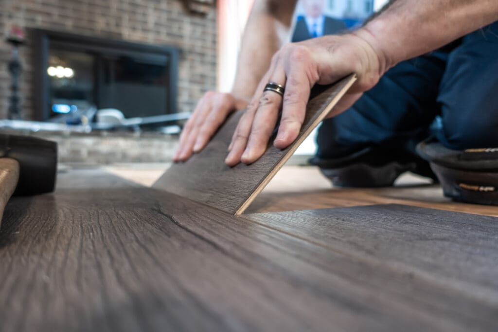 A person skillfully installing wood laminate flooring with expert carpentry precision positions a plank by hand. Behind them, a fireplace and bricks create a cozy atmosphere, reminiscent of an inviting office fit-out that balances functionality and warmth.