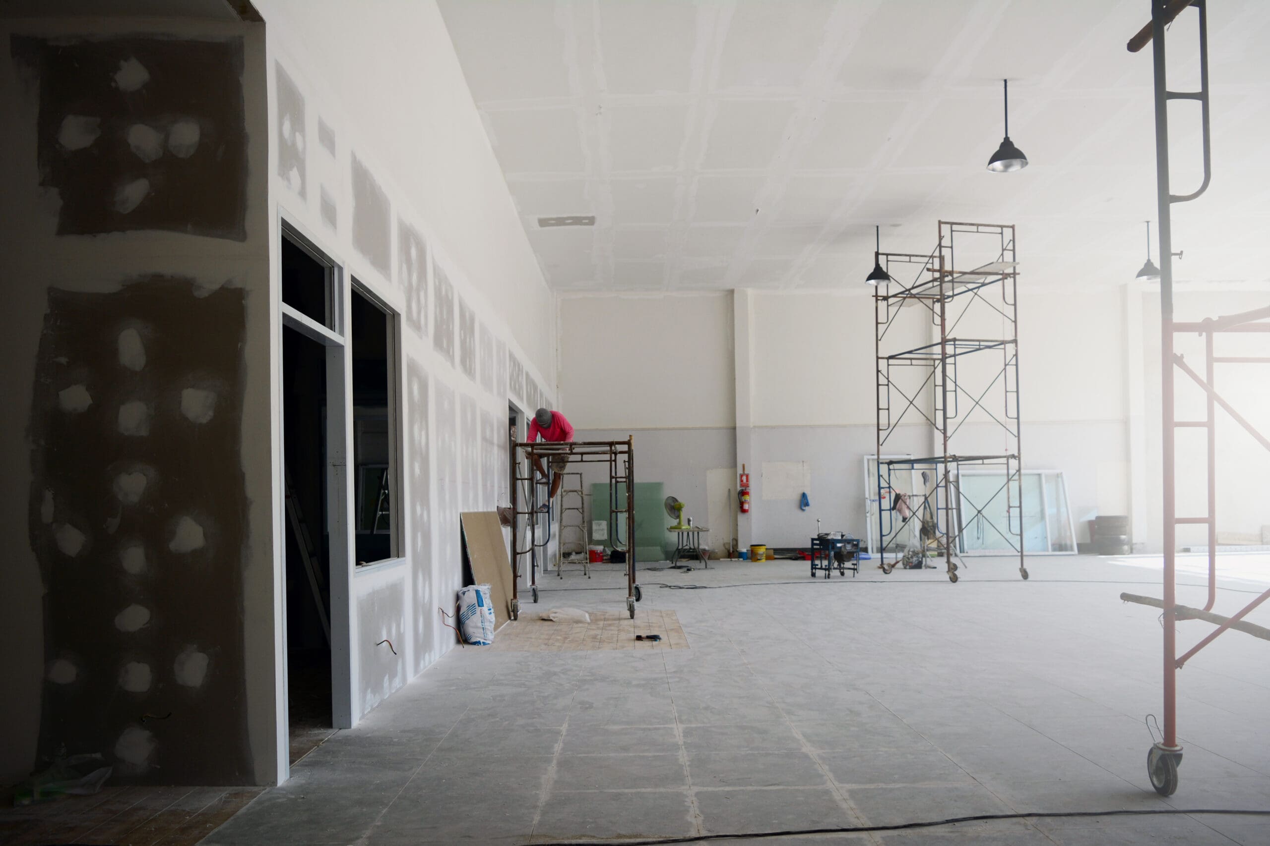 A large, unfinished room with high ceilings hints at an ongoing office fit-out. Scaffolding stands ready as workers paint and perfect the walls. The bare concrete floor is scattered with tools, while drywall partially covers the space, promising transformation under bright overhead lights.