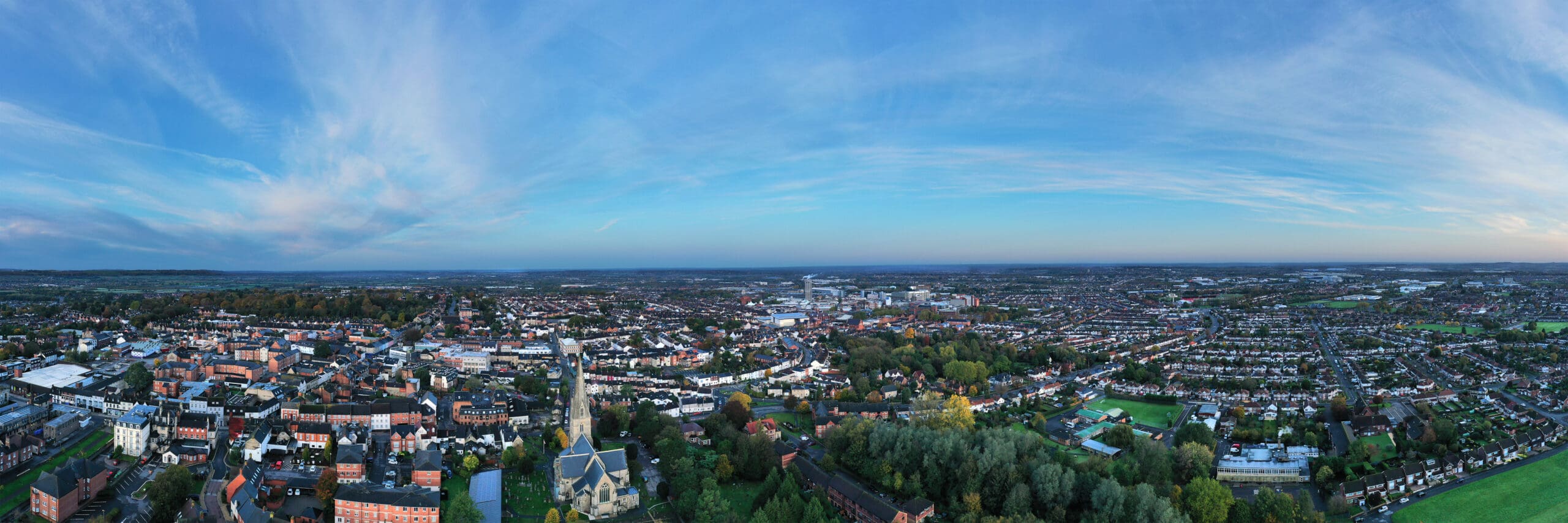 Panoramic view of a sprawling cityscape at sunset. The image captures a mix of urban buildings, greenery, and distant horizons under a blue sky with wispy clouds. A prominent church is visible in the foreground, subtly contrasting the modern office fit-out scattered throughout the city.
