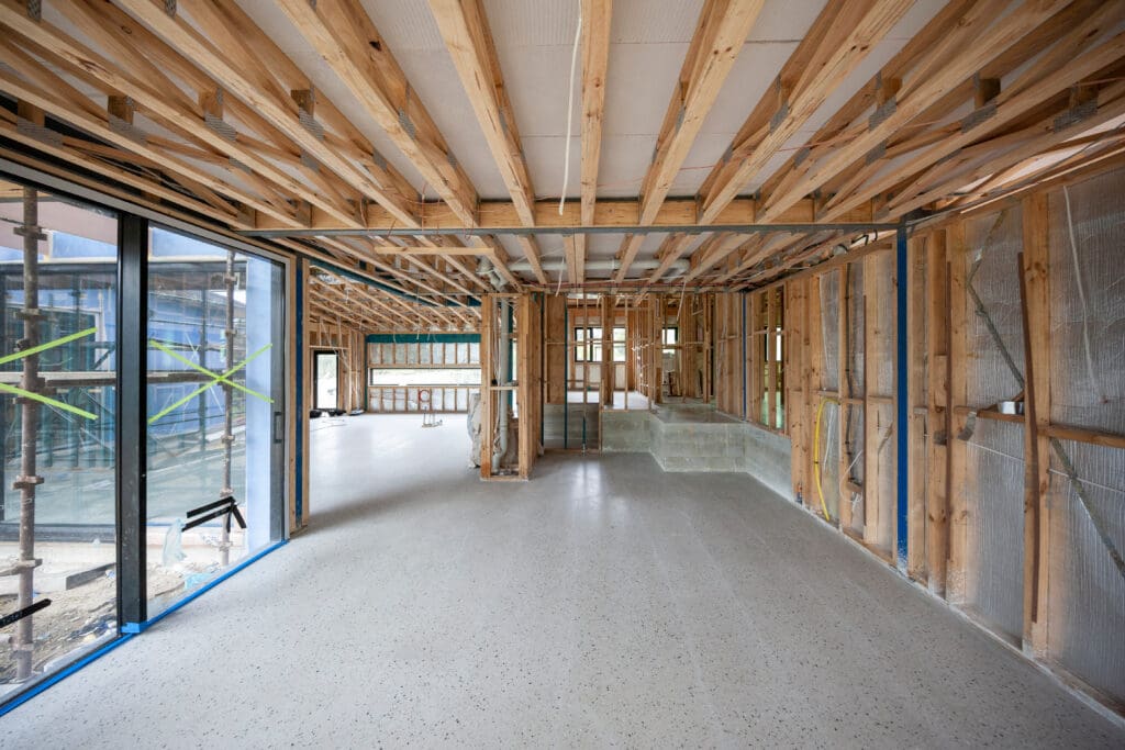 The interior of a building under construction reveals an unfinished room with wooden beams on the ceiling, exposed walls, and large glass windows to the left. A light concrete surface covers the floor as office fit-out progresses. Scaffolding and materials for partitioning are visible outside.