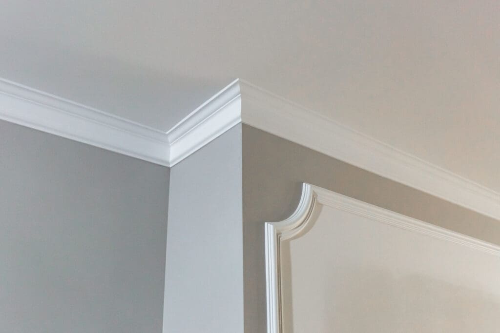 Elegant white crown molding, crafted by skilled carpentry, decorates the corner of a room, contrasting with gray walls and a cream ceiling. The molding features decorative trim, adding a classic architectural detail.