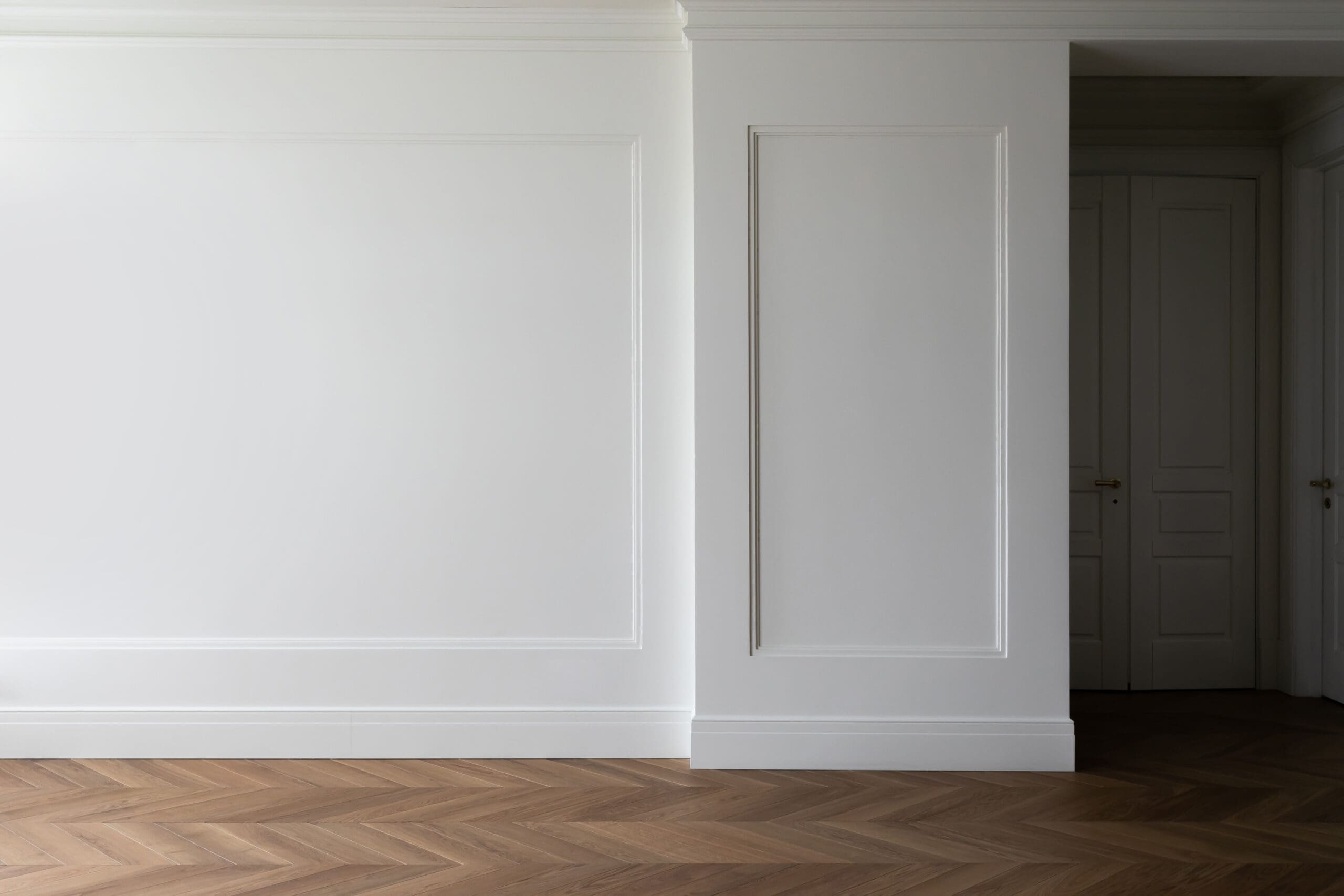 Minimalist interior featuring white paneled walls and herringbone wood flooring. The space, designed with office fit-out elements, includes closed double doors on the right leading to a shadowed area, creating a clean and elegant aesthetic.