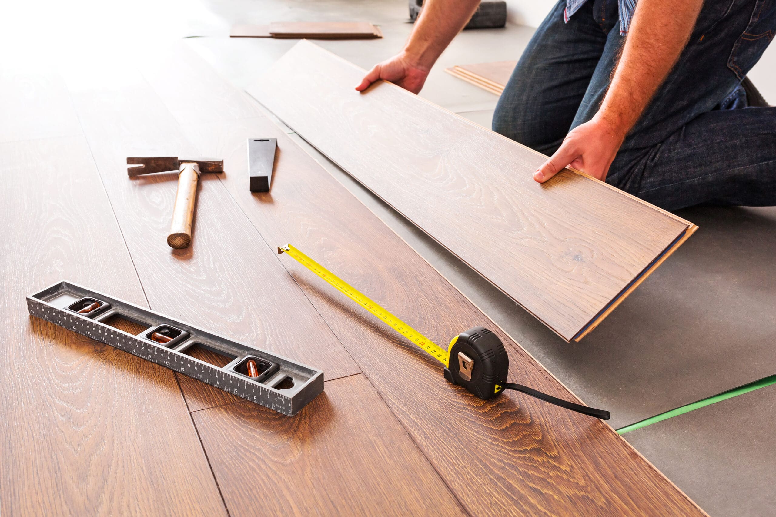 The carpentry expert is meticulously installing a wooden floor, firmly holding a plank. Nearby tools like a hammer, square, measuring tape, and level await use. The floor's light wood grain pattern complements the craftsmanship akin to that of a seasoned dry lining contractor.