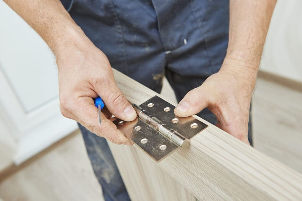 A person in blue work clothes is aligning a metal hinge on the edge of a wooden door, screwdriver in hand. In the background, you see a wooden floor and white wall, typical of an office fit-out setting.