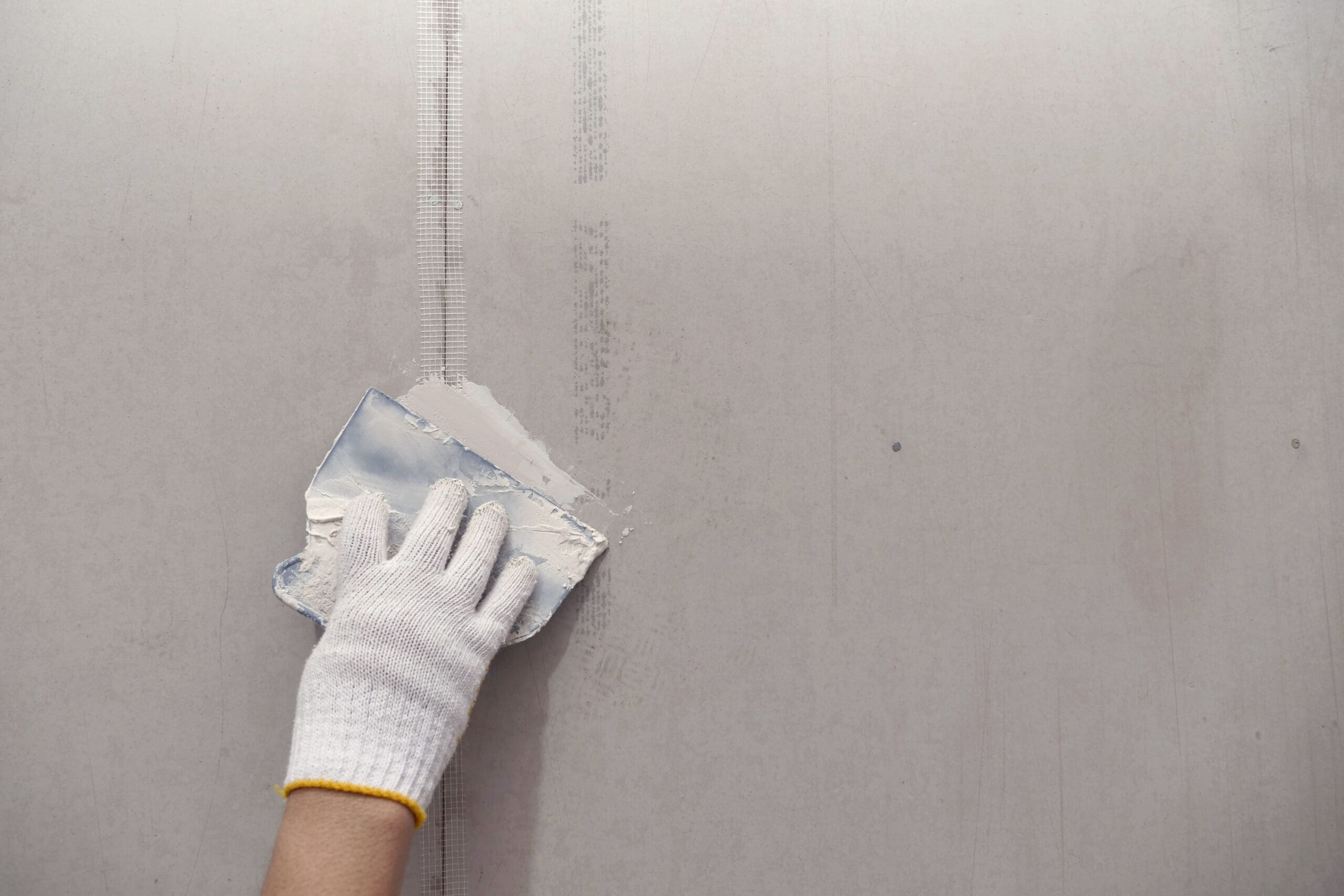 A dry lining contractor wearing a white glove skillfully uses a metal scraper to apply putty on a drywall seam. The plain, unfinished surface reveals visible mesh tape along the seam, highlighting precision often seen in office fit-out projects.