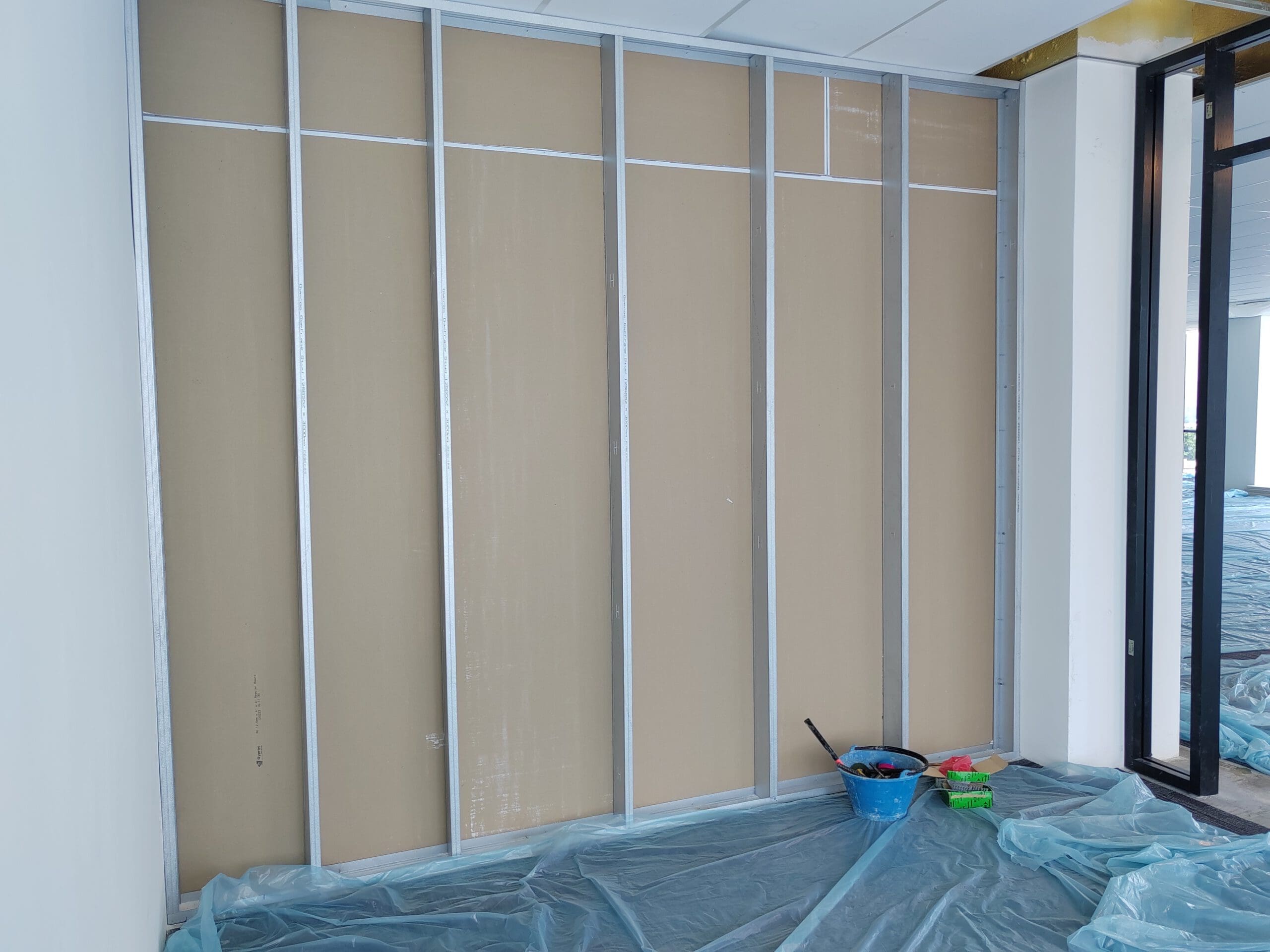 In an unfinished room destined for an office fit-out, a partially constructed drywall with metal studs stands tall. Clear plastic sheeting protects the floor, while a blue bucket and other tools rest nearby, showcasing the precision of a skilled dry lining contractor.