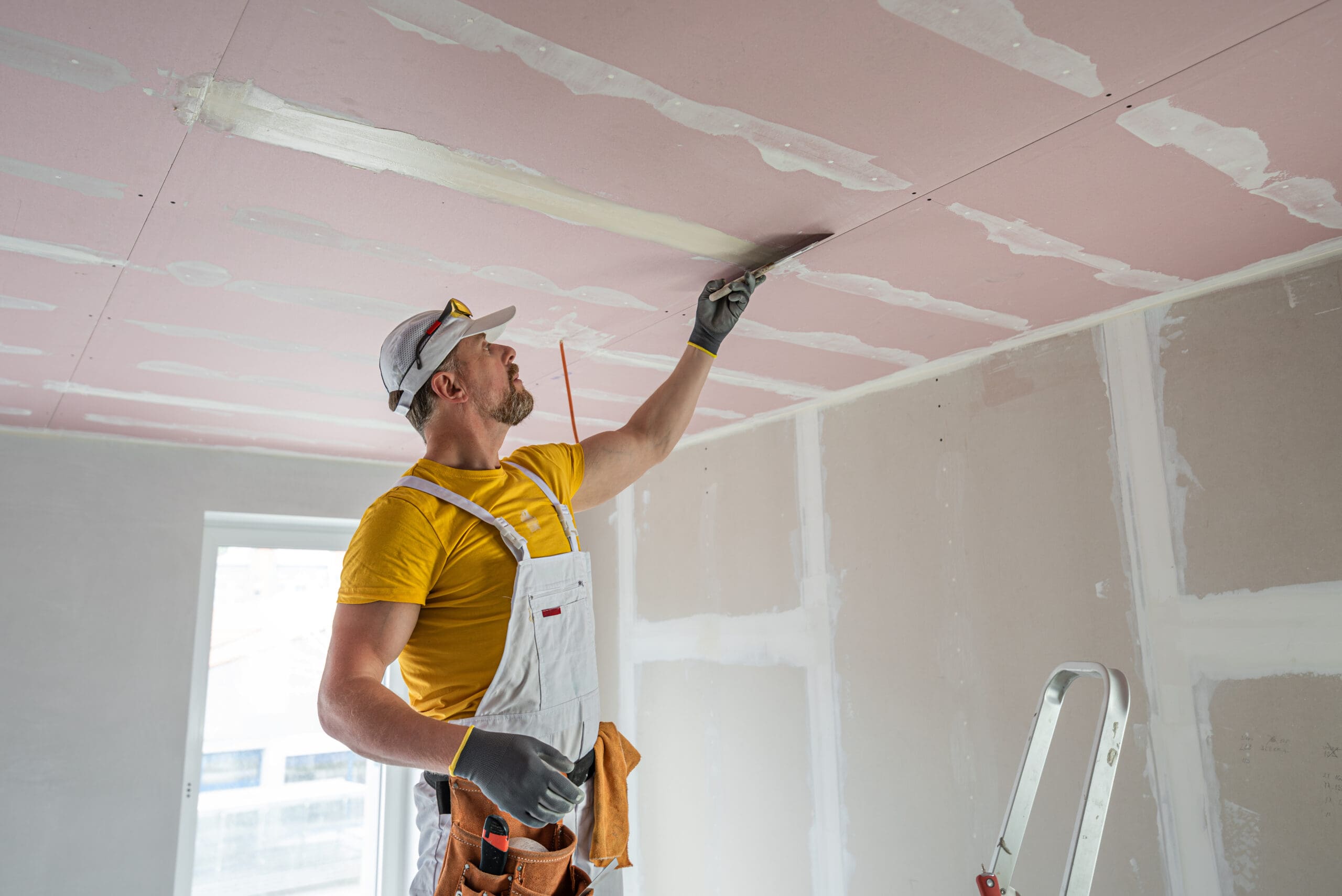 A construction worker in a white cap and overalls is expertly applying joint compound to a suspended ceiling with a trowel. Standing on a ladder, he wears gloves and a yellow shirt in the unfinished room, where partitioning drywall and a window frame the background.