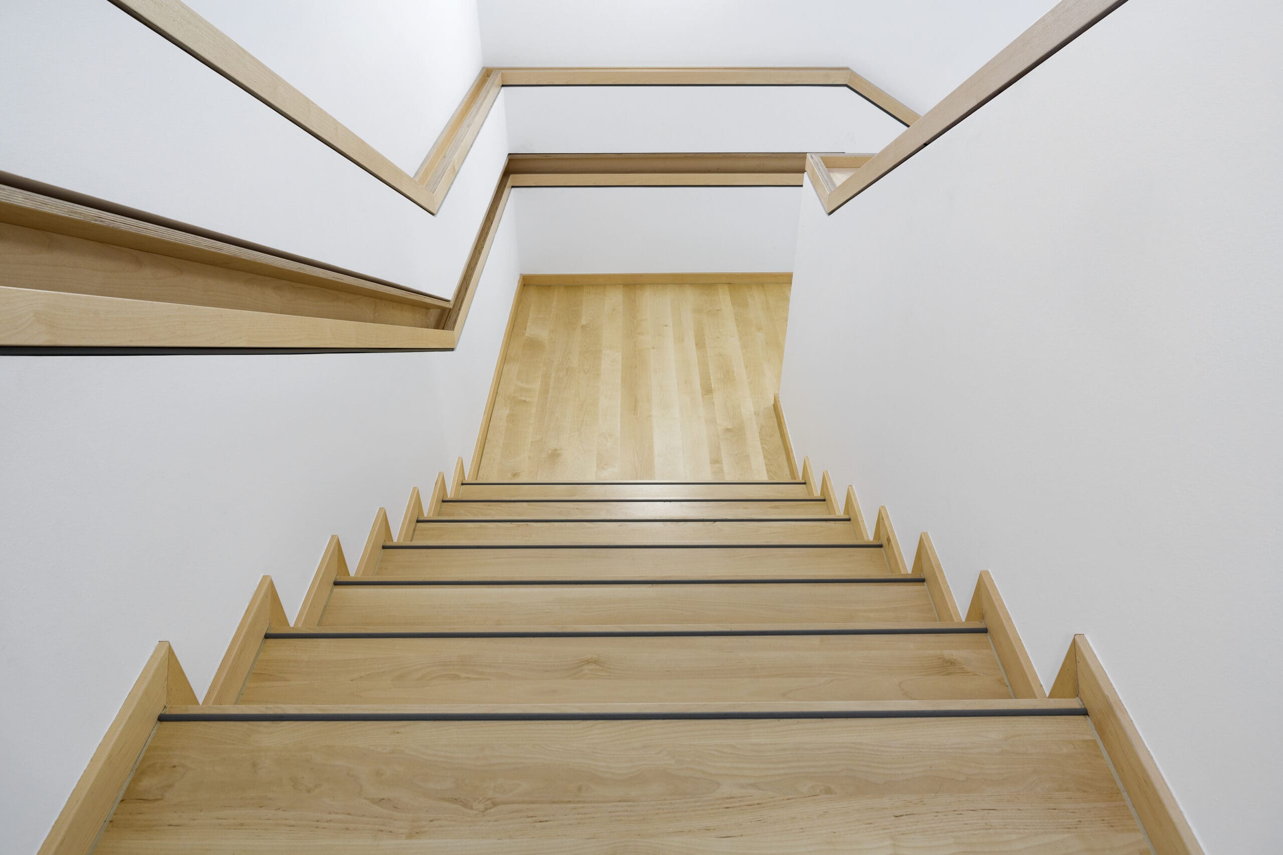 A view from the top of a wooden staircase with light-colored steps and handrails on both sides, reminiscent of an office fit-out. The staircase leads to a landing with matching flooring and continues down, framed by white walls that offer a clean, minimalist aesthetic.