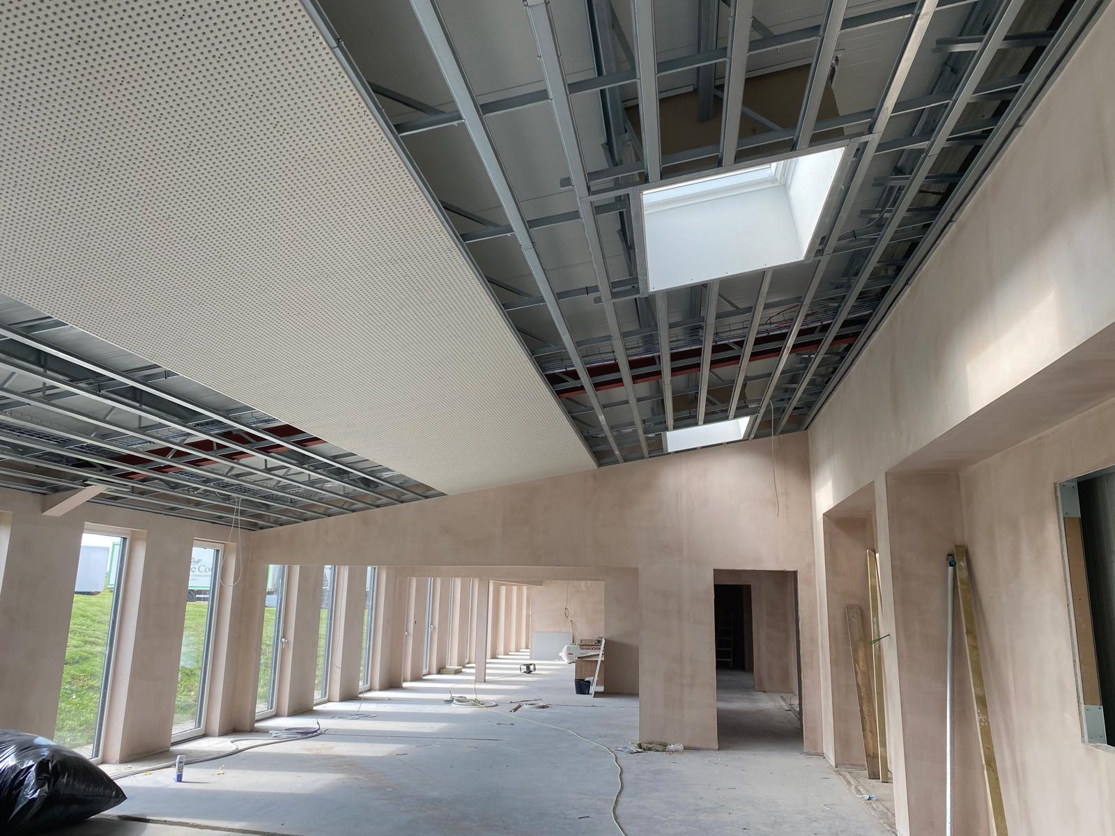 Interior view of a large, unfinished building with visible ceiling framework and drywall. Skylights allow natural light in, illuminating the open space. Construction materials and tools from an office fit-out are scattered on the bare concrete floor.