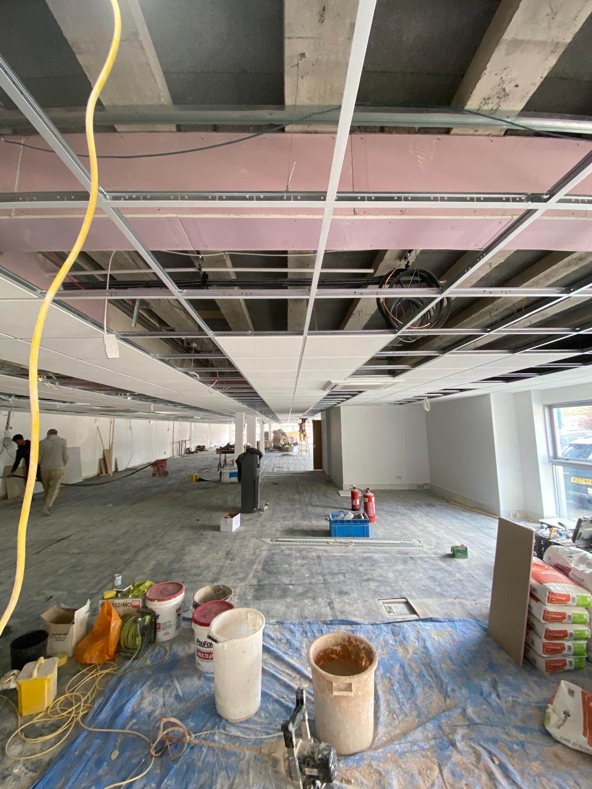 A large indoor construction site features an unfinished ceiling with scattered tools, while a dry lining contractor awaits next steps. Materials like paint buckets, cables, and fire extinguishers are visible. The floor is covered with a tarp, and several workers handle carpentry tasks in the background.