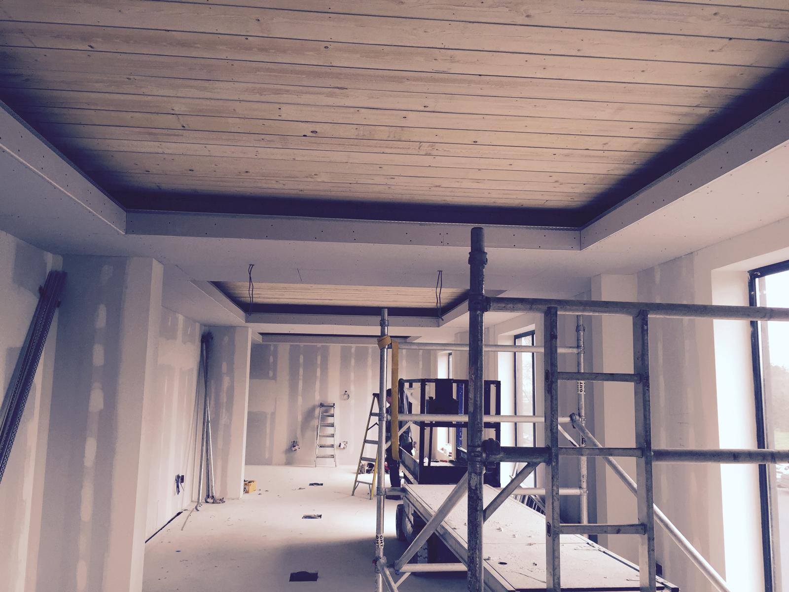 The construction site of a modern building interior buzzes with activity. Unfinished drywall hints at the craftsmanship of a skilled dry lining contractor, while suspended ceilings await installation. Light filters through large windows, casting soft shadows over exposed ceiling beams and scaffolding.