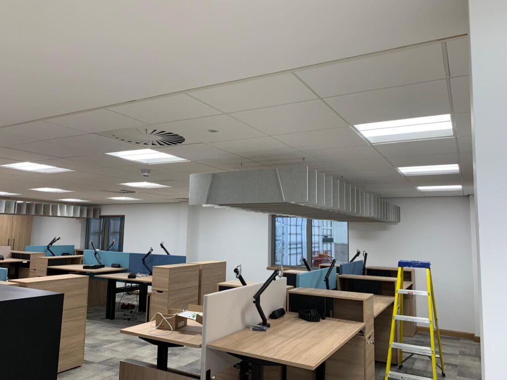 A modern office space features wooden desks and dividers carefully designed through carpentry. Desk lamps are positioned strategically, and acoustic panels hang from the ceiling. A stepladder by the window hints at ongoing office fit-out or maintenance efforts.