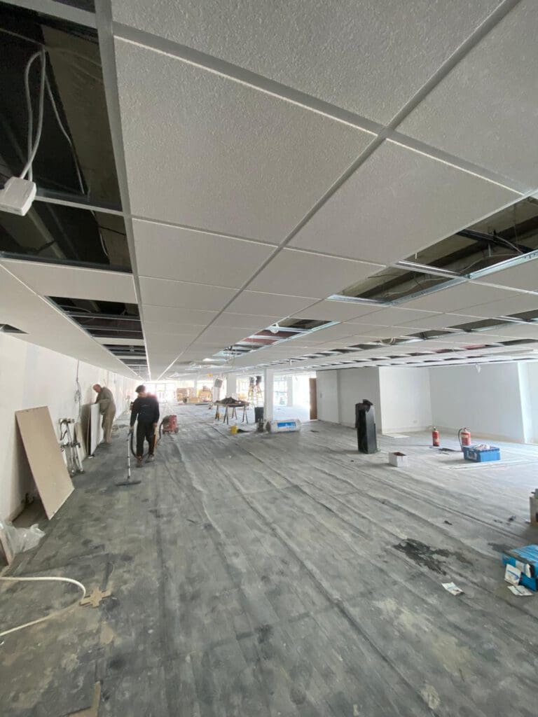 A large room undergoing renovation with workers from a dry lining contractor. The ceiling is partially tiled, and tools and materials are scattered on the floor. One worker stands in the foreground, overseeing partitioning tasks, while others are busy in the background.