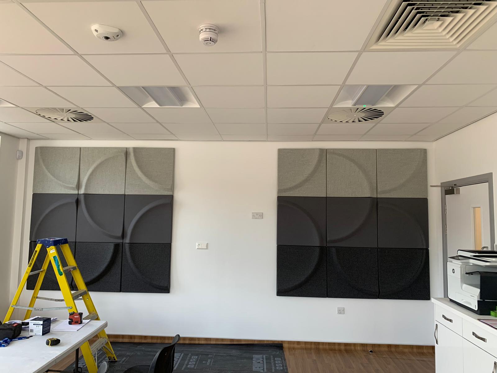 A room showcasing carpentry skills with two square acoustic panels—one light, one dark. On the left, a yellow step ladder and tools suggest an office fit-out in progress. To the right, a printer sits on a countertop beneath suspended ceilings featuring lights and ventilation tiles.