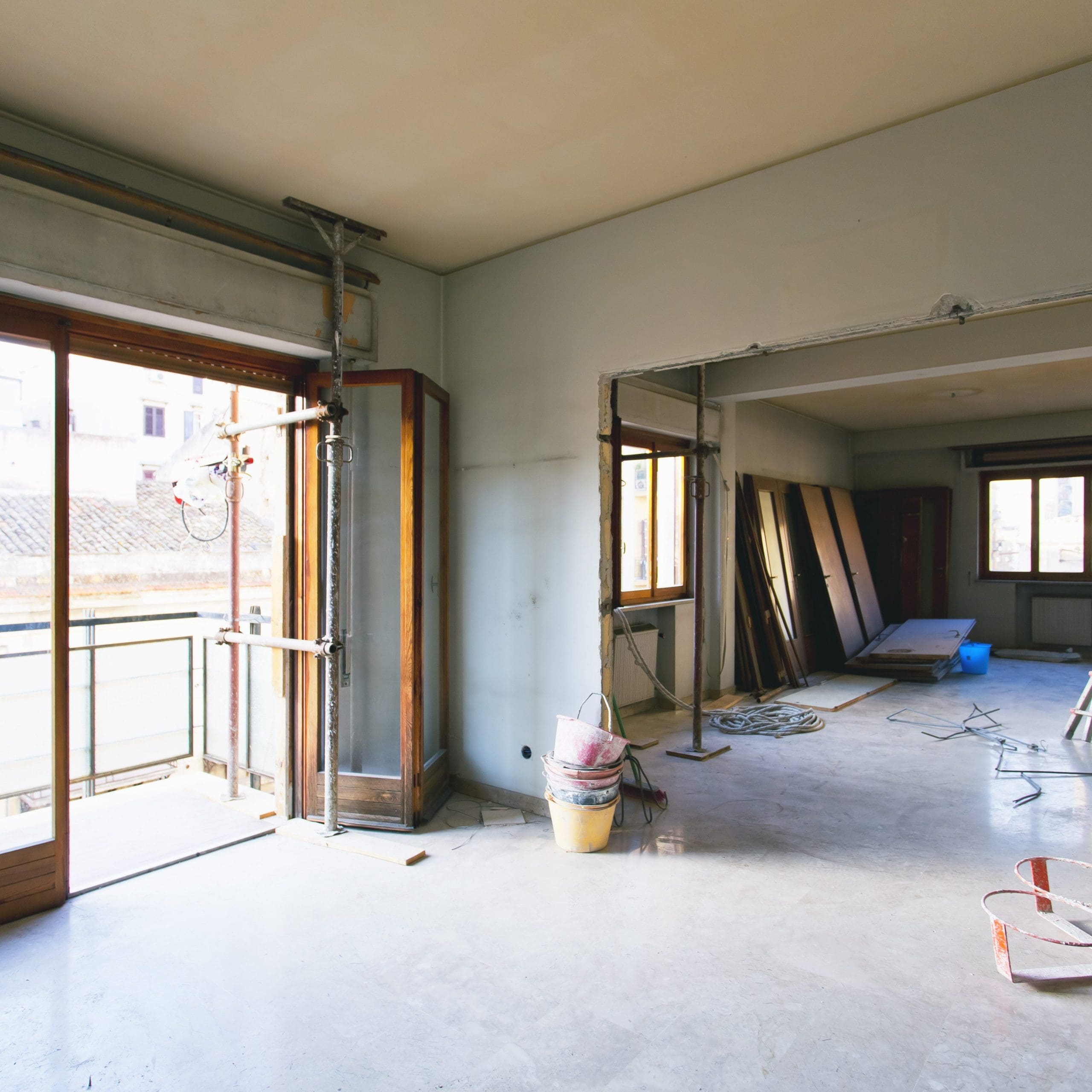 A partially renovated room with light-colored walls and a concrete floor showcases the work of a skilled dry lining contractor. Large windows and a door on the left reveal several wooden planks leaning against the far wall, alongside construction tools essential for carpentry projects.